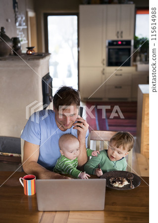 Father sitting at table working on laptop, two sons sitting by him. Older brother eating porridge. Unconditional parental love, family moment. 115614988