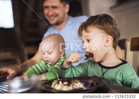 Father sitting at table working on laptop, two sons sitting by him. Older brother eating porridge. Unconditional parental love, family moment. 115615332