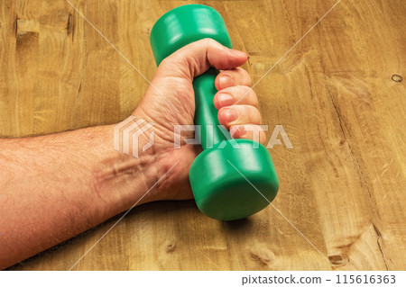 Male hand with dumbbells on a wooden surface. A man's hand holds a green dumbbell against the background of a wooden surface 115616363