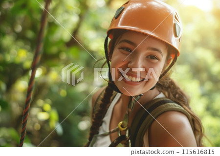 Happy female adventurer with helmet gearing up for zip-line activity in a lush forest 115616815