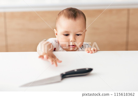 A curious baby, dressed in a white outfit with colorful patterns, reaches for a knife placed on a kitchen counter. The background features wooden cabinets and a light-colored wall 115618362