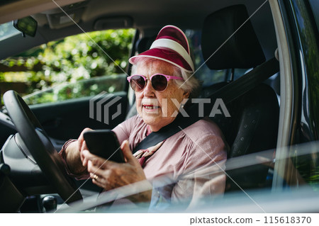 Happy senior woman driving car alone, holding smartphone and scrolling. Safe driving for elderly adults, older driver safety. Happy senior woman driving car alone, holding smartphone and scrolling. Safe driving for elderly adults, older driver safety. 115618370