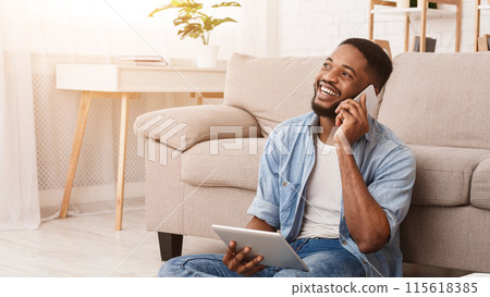 A cheerful African American man is seated comfortably on the floor against a couch, engaged in a pleasant phone conversation, with a tablet in his other hand 115618385