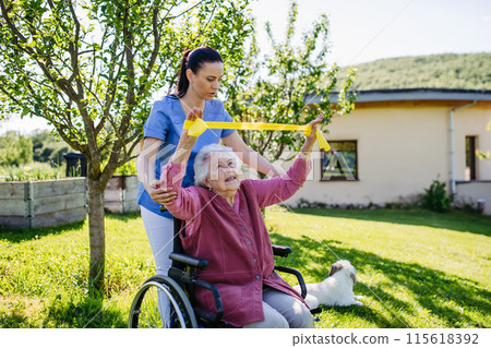 Female caregiver doing motorized exercises with senior woman in wheelchair. Nurse and elderly woman working out with resistance band. 115618392