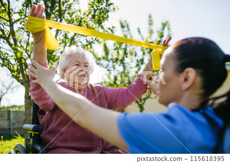 Female caregiver doing motorized exercises with senior woman in wheelchair. Nurse and elderly woman working out with resistance band. Female caregiver doing motorized exercises with senior woman in wheelchair. Nurse and elderly woman working out with resistance band. 115618395