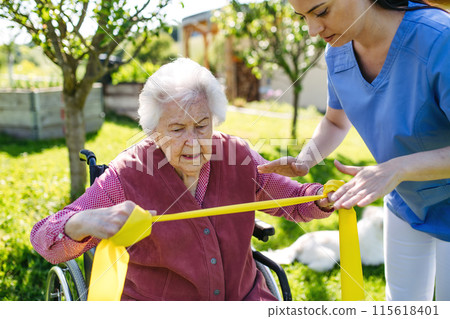 Female caregiver doing motorized exercises with senior woman in wheelchair. Nurse and elderly woman working out with resistance band. Female caregiver doing motorized exercises with senior woman in wheelchair. Nurse and elderly woman working out with resistance band. 115618401