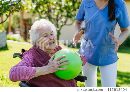 Female caregiver doing motorized exercises with senior woman in wheelchair. Nurse and elderly woman working out with exercise ball. Female caregiver doing motorized exercises with senior woman in wheelchair. Nurse and elderly woman working out with exercise ball. 115618404