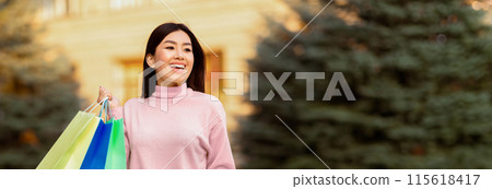 Asian woman is standing outdoors holding multiple shopping bags in her hands. She appears to have just finished shopping and is carrying her purchases. The bags are colorful and seem full, panorama Asian woman is standing outdoors holding multiple shopping bags in her hands. She appears to have just finished shopping and is carrying her purchases. The bags are colorful and seem full, panorama 115618417