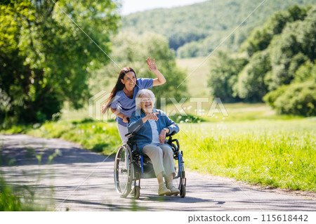 Female caregiver pushing senior woman in wheelchair. Nurse and elderly woman enjoying a warm day in nursing home, public park. Female caregiver pushing senior woman in wheelchair. Nurse and elderly woman enjoying a warm day in nursing home, public park. 115618442