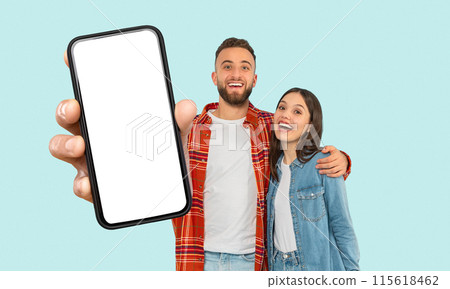 A happy couple is standing together in front of a blue background. The man is holding a black smartphone with a white screen in his right hand. The couple is smiling and looking at the camera. 115618462