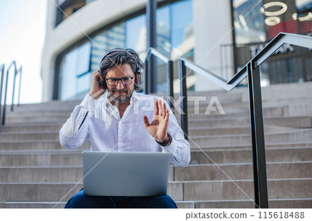 Portrait of mature businessman sitting on stairs in city, working on laptop. Handsome man with glasses in suit. 115618488