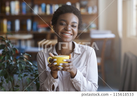 Out of home breakfast. Smiling beautiful African American millennial woman wearing smart casual outfit having coffee in cafe, holding yellow mug and smiling at camera, copy space Out of home breakfast. Smiling beautiful African American millennial woman wearing smart casual outfit having coffee in cafe, holding yellow mug and smiling at camera, copy space 115618533