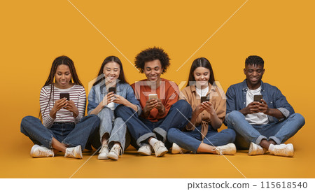 This image features a diverse group of five young adults, all smiling and looking down at their smartphones. They are sitting cross-legged on a yellow background This image features a diverse group of five young adults, all smiling and looking down at their smartphones. They are sitting cross-legged on a yellow background 115618540