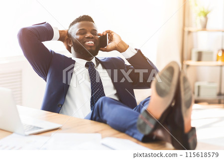 Positive african american manager talking by phone while having break at office, leaning back on chair and putting legs on table, empty space Positive african american manager talking by phone while having break at office, leaning back on chair and putting legs on table, empty space 115618579