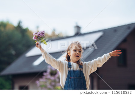 Beautiful young girl with purple lilacs flower in hands. Young girl spending time outdoors in nature, springtime. House with solar panels in background. Beautiful young girl with purple lilacs flower in hands. Young girl spending time outdoors in nature, springtime. House with solar panels in background. 115618582