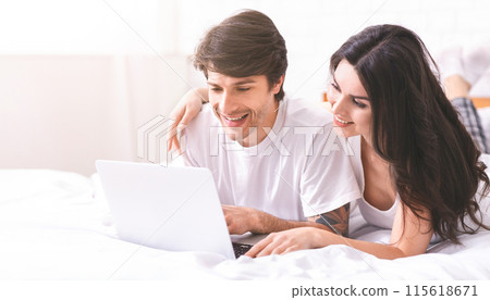 A young couple is lying in bed, using a laptop computer. The man is looking at the screen with a smile on his face while the woman is looking at him, also smiling 115618671