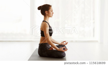 A woman is sitting cross-legged on the floor, practicing yoga. She is focused and holding a pose with her eyes closed, showing concentration and mindfulness, copy space 115618699