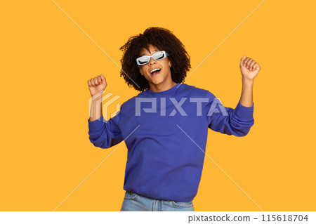 A joyful young guy with curly hair and sunglasses dances with hands raised against a solid yellow background, wearing a blue sweatshirt and jeans, expressing happiness and carefree energy. 115618704