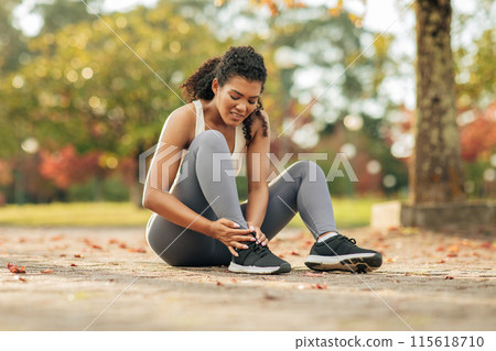 A young woman wearing athletic attire is adjusting her running shoes while sitting on a path in an autumn park. Lady runner injured ankle 115618710