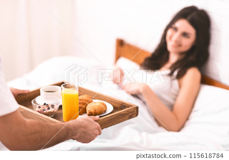 A man is bringing a breakfast tray to a woman in bed. The tray contains a cup of coffee, a glass of orange juice, a bowl of cereal, and two croissants. The woman is smiling and looking at the tray. A man is bringing a breakfast tray to a woman in bed. The tray contains a cup of coffee, a glass of orange juice, a bowl of cereal, and two croissants. The woman is smiling and looking at the tray. 115618784