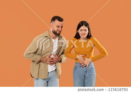 A young couple holds their stomachs, appearing to endure discomfort against an orange backdrop. Both are casually dressed, emphasizing their distress. 115618793