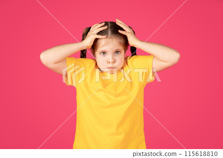 Confused Little Girl Making Funny Face And Touching Head With Hands, Preteen Female Child In Yellow T-Shirt Fooling And Looking At Camera, Standing Isolated Over Pink Studio Background, Copy Space 115618810