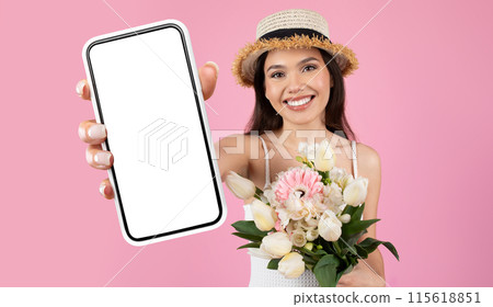 A young woman wearing a straw hat smiles at the camera as she holds a white smartphone with a blank screen and a bouquet of white and pink flowers. The image is shot against a solid pink background. A young woman wearing a straw hat smiles at the camera as she holds a white smartphone with a blank screen and a bouquet of white and pink flowers. The image is shot against a solid pink background. 115618851