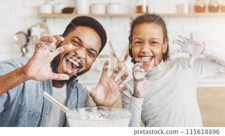 Happy african dad and daughter having fun while cooking pastry, making funny growling faces and imitating animal paws, kitchen interior Happy african dad and daughter having fun while cooking pastry, making funny growling faces and imitating animal paws, kitchen interior 115618896