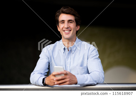 Portrait of cheerful handsome young man in formal outfit using smartphone while standing at office building balcony, having break. Modern technologies in business, communication concept Portrait of cheerful handsome young man in formal outfit using smartphone while standing at office building balcony, having break. Modern technologies in business, communication concept 115618907