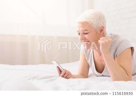 Good news. Happy senior woman lying on bed, smiling and reading text message on cellphone, enjoying win, panorama with empty space Good news. Happy senior woman lying on bed, smiling and reading text message on cellphone, enjoying win, panorama with empty space 115618930