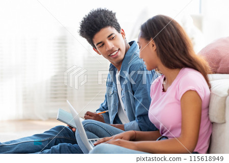 African American teen couple sits on the floor, the guy is working on a laptop while the girl is writing in a notepad. They are engaged in a conversation, the guy seems to be explaining something 115618949