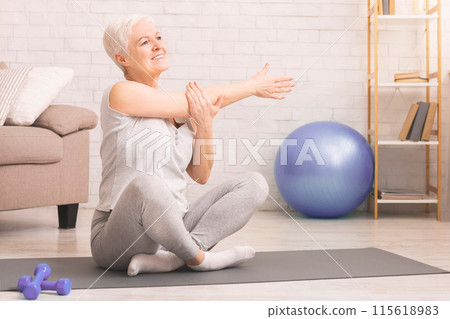 Senior woman is seated on a yoga mat in a cozy living room. She appears focused and engaged in a yoga practice, stretching arms 115618983