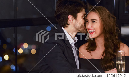 Loving couple on date in luxury restaurant. Man whispering into girlfriends ear. Love and romance concept Loving couple on date in luxury restaurant. Man whispering into girlfriends ear. Love and romance concept 115619042