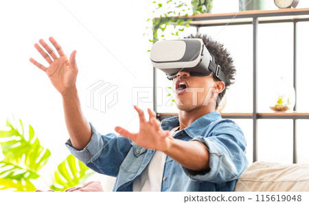 African American teen guy wearing a virtual reality headset sits on a couch, his hands outstretched, and his mouth open in surprise. He appears to be immersed in a virtual reality experience 115619048