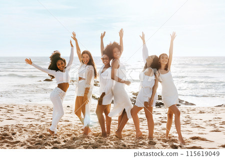Happy multiracial ladies dancing with arms raised while celebrating hen party on the beach at sunny day, full length. Joyful young women bride's team Happy multiracial ladies dancing with arms raised while celebrating hen party on the beach at sunny day, full length. Joyful young women bride's team 115619049