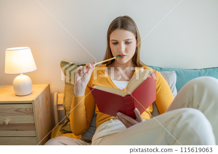 A girl is seated on a bed, engrossed in a book she holds in her hands. The room is well-lit, and she appears focused on the text as she reads. 115619063
