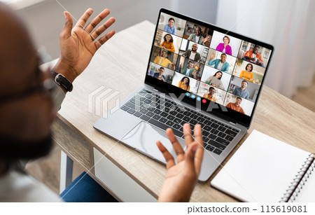 Black man sits at a desk in front of a laptop screen, participating in a video conference meeting. He has both hands raised, gesturing to the other participants in the call. 115619081