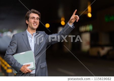 Happy handsome young businessman wearing formal outfit raising han up, catching taxi on the street, holding laptop and smartphone, entrepreneur going to business meeting, copy space 115619082