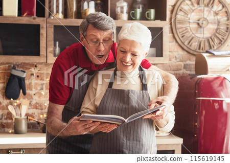 A senior couple, wearing aprons, stands together in a modern kitchen, smiling as they read a recipe book. The man is standing behind the woman, who is holding the book open. A senior couple, wearing aprons, stands together in a modern kitchen, smiling as they read a recipe book. The man is standing behind the woman, who is holding the book open. 115619145