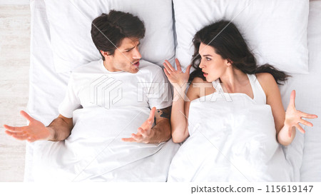 A young couple is lying in bed, arguing with each other. The man is lying on his back with his arms outstretched, while the woman is lying on her side, facing him. They both appear to be angry A young couple is lying in bed, arguing with each other. The man is lying on his back with his arms outstretched, while the woman is lying on her side, facing him. They both appear to be angry 115619147