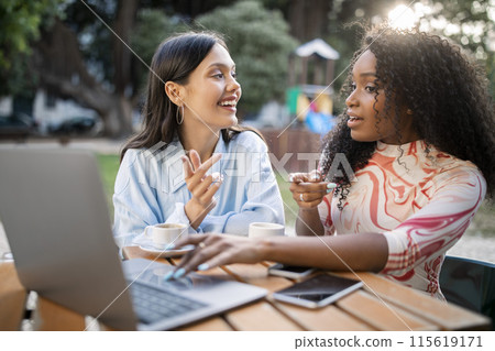 Cheerful Female Friends Shopping Online On Laptop While Resting In Cafe Outdoors, Two Happy Multiethnic Women Pointing At Screen And Discussing Purchases, Sitting At Table Outside And Drinking Coffee 115619171