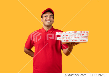 Delivery man wearing a red shirt is standing while holding a pizza box, posing isolated on yellow studio background Delivery man wearing a red shirt is standing while holding a pizza box, posing isolated on yellow studio background 115619230