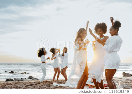 Group of carefree young women toasting wine glasses while dancing and having fun on the beach, having hen party at coastline. Female friends enjoying their vacation Group of carefree young women toasting wine glasses while dancing and having fun on the beach, having hen party at coastline. Female friends enjoying their vacation 115619249