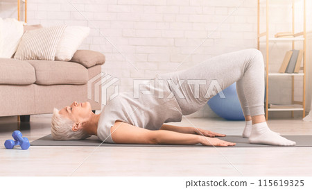 A content senior woman with short hair lies on her back carrying out a bridge exercise on a yoga mat in spacious, well-lit living room. She wears comfortable attire and has pair of dumbbells nearby A content senior woman with short hair lies on her back carrying out a bridge exercise on a yoga mat in spacious, well-lit living room. She wears comfortable attire and has pair of dumbbells nearby 115619325