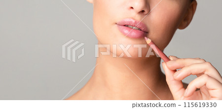 A close-up of a woman face as she carefully applies a pink lip liner to her lips, demonstrating a makeup technique. The background is plain and neutral, cropped, copy space 115619330