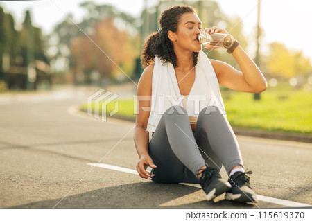 A woman is seated on the side of a road, holding a bottle to her mouth and drinking from it. She appears relaxed and focused on her drink. A woman is seated on the side of a road, holding a bottle to her mouth and drinking from it. She appears relaxed and focused on her drink. 115619370
