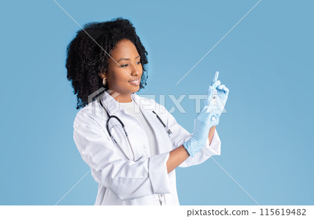 Cheerful young black lady doctor in white coat, protective gloves shows syringe, isolated on blue studio background. Disease treatment, health care, vaccination medicine in clinic Cheerful young black lady doctor in white coat, protective gloves shows syringe, isolated on blue studio background. Disease treatment, health care, vaccination medicine in clinic 115619482