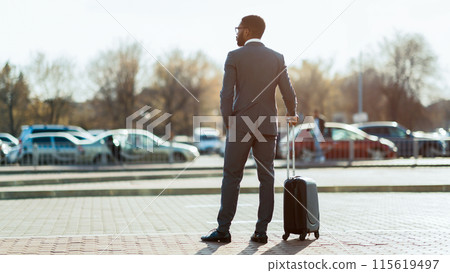 African American businessman wearing suit with suitcase crossing street, going vacation, back view, looking at copy space, panorama African American businessman wearing suit with suitcase crossing street, going vacation, back view, looking at copy space, panorama 115619497