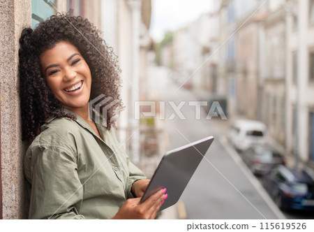 Cheerful Black Woman Holding Digital Tablet Standing At Balcony Of Her Apartment With Street View, Smiling To Camera While Browsing Internet Spending Time Online. Computer Gadgets, Technology 115619526