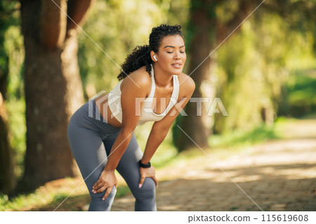 A woman in athletic wear is resting during her jog on a sunlit forest trail. She is leaning forward with hands on knees, wearing wireless earbuds, and looks relaxed in the peaceful surroundings. 115619608